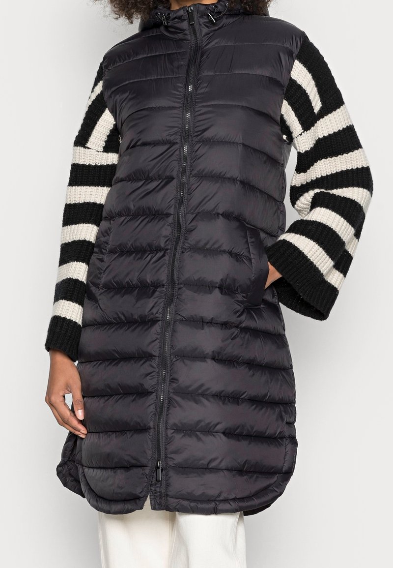 Personne portant un gilet matelassé noir sur un pull en tricot rayé noir et blanc à manches larges, main dans la poche.