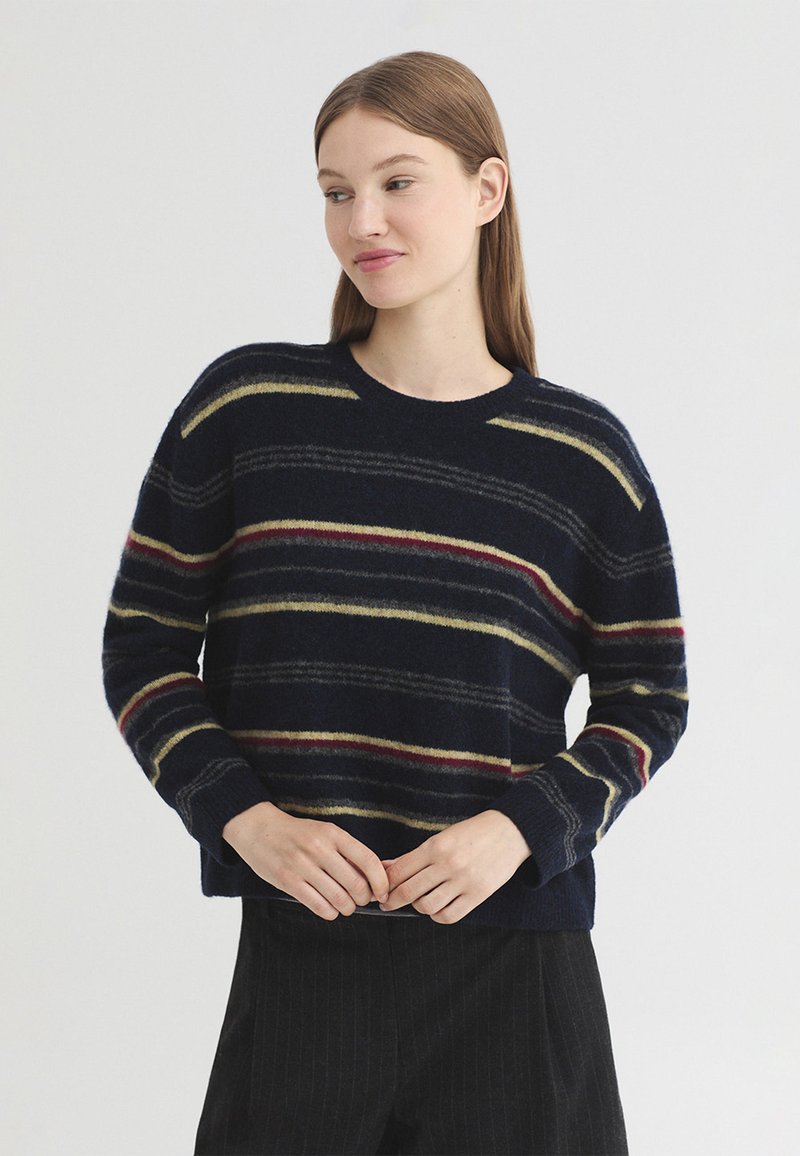 Femme aux cheveux bruns et raides portant un pull foncé avec des rayures horizontales beige, rouge et gris, et un pantalon sombre, debout devant un fond uni.