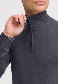 Pull-over gris avec un col à côtes hautes et une fermeture éclair quart de tour. Fabriqué en tissu tricoté lisse, avec des manches longues et une silhouette ajustée.