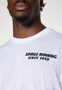 Bijela pamučna majica s okruglim izrezom, koja na lijevoj strani prsa ima crni grafički natpis "ADIDAS RUNNING SINCE 1949".