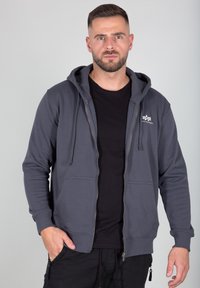 Sudadera gris con cremallera, con un bolsillo frontal, capucha con cordones y puños acanalados. Llevada sobre una camiseta negra, exhibida contra un fondo neutro.