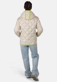 Giacca puffer in crema trapuntata con cappuccio, caratterizzata da un accento verde lungo il colletto e l'orlo, abbinata a jeans azzurri chiari e sneakers grigie.