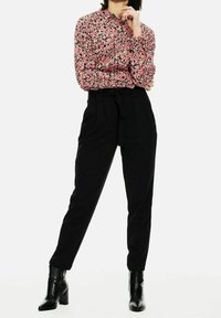 Bloemige blouse met lange mouwen in roze en zwart met een knoopsluiting, gecombineerd met zwarte, high-waisted broeken. Model draagt zwarte hakken.