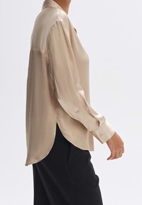 Blouse en satin beige avec col, manches longues et ourlet arrondi, présentant une texture lisse et des fermetures par boutons discrets.
