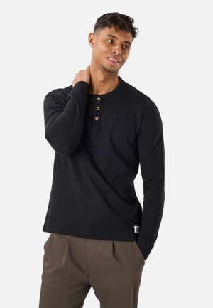 Młody mężczyzna w czarnej koszulce henley z długim rękawem i brązowych spodniach, z jedną ręką dotykającą szyi, na jednolitym tle.