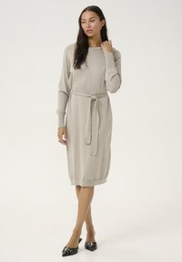 Robe en tricot beige à manches longues avec une ceinture assortie, encolure ronde, poignets côtelés et ourlet, accompagnée de ballerines noires pointues.