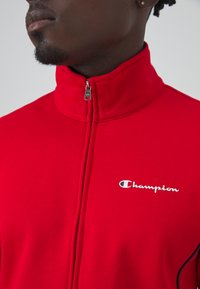 Chaqueta roja con cremallera fabricada en un material suave, con un cuello alto y un logo blanco de "Champion" en el lado izquierdo.