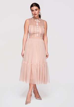 Frau trägt ein ärmelloses Midi-Kleid in zartem Rosa mit Spitzen-Details, Schleife am Kragen und transparentem Überwurf, kombiniert mit silbernen spitzen Absätzen.