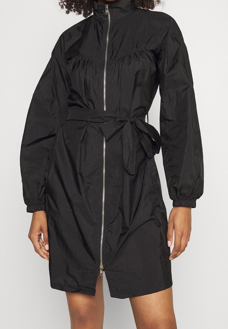 Robe noire en tissu léger, avec col montant, fermeture éclair à l'avant, poignets élastiques et ceinture nouée à la taille. Texture lisse.