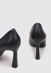 Chaussures à talons hauts noires avec une finition en cuir lisse, un design à bout pointu et des talons minces et angulaires. Style minimaliste sans ornementations.