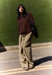 Brown fuzzy sweater, beige wide-leg trousers, black handbag, and brown footwear. Background features green tiled wall and paved street.