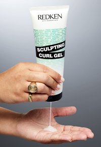 Redken Sculpting Curl Gel in een witte tube met turquoise en zwarte tekst, die heldere gel op een hand dispenseert. Tekst beschrijft verschillende voordelen voor het haar.