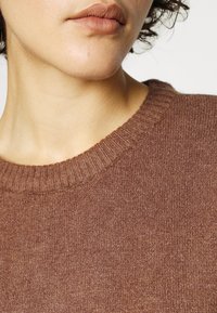 Pull marron en tricot avec un col rond côtelé, présentant une texture douce et des variations subtiles de couleur sur le tissu.