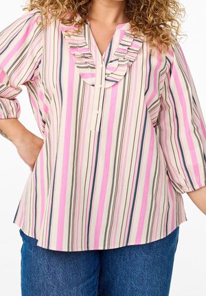 Vrouw draagt een blouse met verticale strepen in roze, beige en zwart, met een ruches kraag en driekwart mouwen, gecombineerd met een blauwe spijkerbroek.