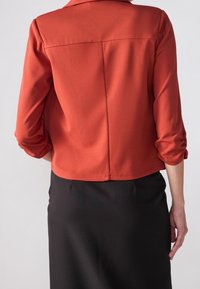 Chaqueta corta de color óxido hecha de una tela suave con mangas de tres cuartos, que presenta una costura trasera limpia y un dobladillo oculto. Combinada con una falda negra.