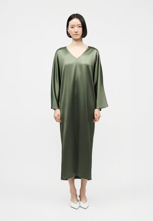 Γυναίκα που στέκεται φορώντας ένα μακρύ, φαρδύ, ελιάς πράσινο σατέν φόρεμα με μακριά μανίκια, σετ με λευκές μυτερές γόβες, μπροστά σε απλό φόντο.