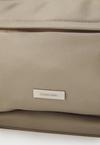 Beżowa torba nylonowa o gładkiej fakturze, z poziomym zapięciem na zamek błyskawiczny i srebrną tabliczką z nazwą Calvin Klein z przodu.