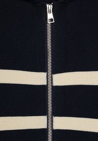Námornícky zip-up svetr s dvěma vodorovnými krémovými pruhy, texturovaným pleteným materiálem a středovým zipem z nerezové oceli.