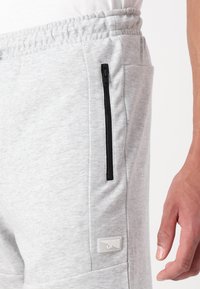 Lysgrå joggebukser laget av mykt stoff, med en svart glidelåslomme på siden og et hvitt logo-påskrift nær midjen.