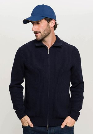 Marineblauer, gerippter Strick-Reißverschluss-Pullover mit hohem Kragen und langen Ärmeln, kombiniert mit einer blauen Mütze. Der Stoff hat eine dicke und warme Textur.