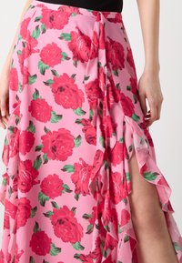 Jupe florale rose ornée de grandes roses rouges et de feuilles vertes, avec des bords ondulés et une fente sur le côté pour un détail supplémentaire. Fabriquée en tissu léger.