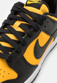 Svarta och gula sneakers med överdel i läder, texturerade svarta skosnören och en vit mellansula med en framträdande Nike-logga i svart.