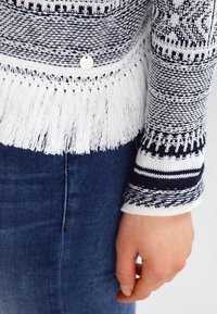 Person som bär mörkblå jeans och en svart-vit mönstrad stickad tröja med vit frans i fållen, med handen avslappnat vid sidan.