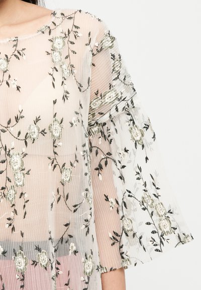 Surimpression transparente brodée de fleurs en beige clair et vert, avec une texture côtelée et des manches évasées. Accents délicats sur l'ensemble.