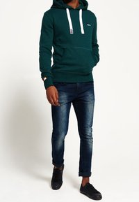 Sudadera verde con cordones blancos y bolsillo frontal, combinada con jeans oscuros. Modelo lleva zapatillas negras. Diseño y textura simples.