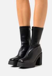 Bottes noires à cheville en cuir lisse, dotées d'une fermeture éclair, d'un talon haut texturé et d'un bout arrondi.