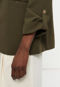 Sötétebb bőrű kar olívazöld, gombbal ellátott hosszú ujjú felsőben, fehér nadrággal világos háttér előtt.