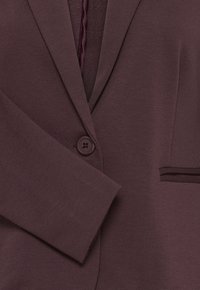 Blazer marron foncé à un bouton avec revers crantés et une poche passepoilée sur le côté droit, vu de face.