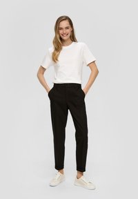 T-shirt blanc à rayures légères, pantalon noir ajusté et baskets beiges. Le modèle se tient avec les mains dans les poches sur un fond neutre.