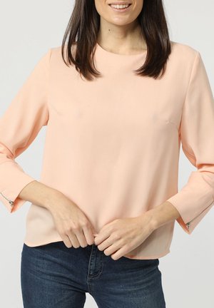 Blusa de manga larga en color melocotón claro, con cuello redondo y puños con cremallera. La tela parece suave y ligera, diseñada para un ajuste relajado.