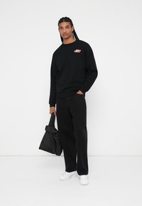 Sudadera negra con un pequeño logo colorido, combinada con pantalones anchos negros y zapatillas blancas. Sosteniendo un bolso tote negro acolchado.