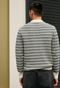Maglione a righe in nero e crema, tessuto strutturato, scollatura rotonda, polsini e orlo a coste, abbinato a jeans scuri, visto da dietro.