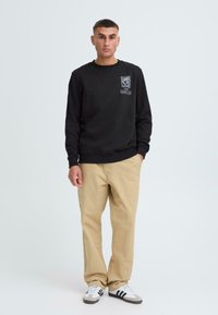 Black crewneck sweatshirt features a graphic logo on the chest. Paired with beige straight-leg pants and gray and white sneakers.
