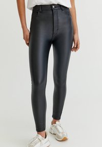 Leggings de imitación de cuero negros de talle alto con un ajuste ceñido, textura suave y cierre de cremallera, combinados con zapatillas blancas y beige.