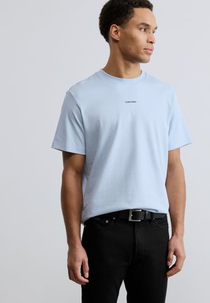 Tricou albastru deschis din bumbac cu mâneci scurte, având un mic logo negru „Calvin Klein” centrat pe piept, asortat cu blugi negri.