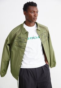 Ελαφρύ μπουφάν ελαιολάδου με διχτυωτές λεπτομέρειες, σε συνδυασμό με λευκό T-shirt που φέρει το πράσινο λογότυπο Jordan και μαύρο παντελόνι.