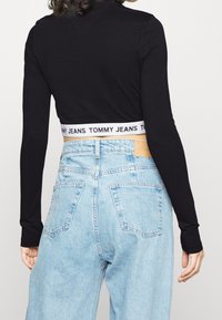 Black long-sleeve crop top with a white branded elastic band. Worn with light blue high-waisted jeans featuring multiple pockets.