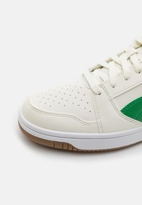 Weiße synthetische Sneaker mit perforierter Zehenpartie, grünem Akzentpanel, strukturierten Seiten, beiger Gummisohle und weißen flachen Schnürsenkeln.