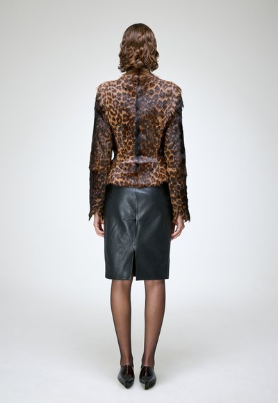 Femme aux cheveux bruns ondulés portant une veste en fourrure à imprimé léopard, une jupe crayon en cuir noir, des collants noirs transparents et des chaussures noires pointues, vue de dos.