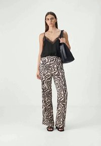 Camisole noir avec bordure en dentelle, associé à un pantalon large à imprimé zèbre marron et crème. Le modèle tient un sac à main noir et porte des chaussures noires.