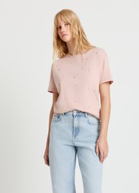 Hellrosa T-Shirt aus Baumwolle mit kurzen Ärmeln, das vorne gleichmäßig mit kleinen dekorativen Strasssteinen besetzt ist. Getragen zu hoch taillierten blauen Jeans.