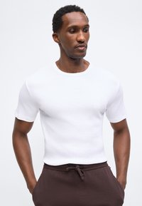 Homme aux cheveux courts et bouclés portant une chemise blanche ajustée à manches courtes et un pantalon marron foncé texturé, les mains dans les poches.