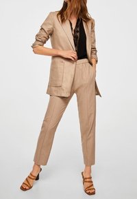 Blazer beige cintré avec deux poches avant, associé à un pantalon fuselé assorti. Le modèle porte un haut noir et des sandales à talons marron.