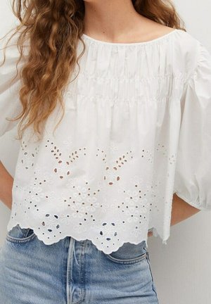 Vrouw draagt een witte blouse met opengewerkte bloemenborduursels en pofmouwen, gecombineerd met blauwe spijkerbroeken.