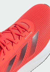 Chaussure de course rouge avec tige en mesh, fermeture à lacets, bandes noires inclinées sur le côté et semelle blanche amortissante.