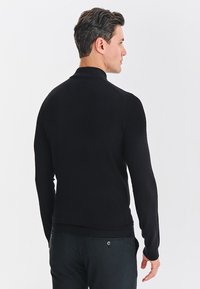 Czarny dziany sweter z wysokim kołnierzem i dopasowaną sylwetką. Długie rękawy, prążkowany dół oraz teksturowana tkanina. Noszony z ciemnymi spodniami.